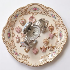 May include: A silver-toned watch with a hinged cover, set on a decorative, floral-patterned plate. The watch face is surrounded by small crystals. The plate is adorned with heart-shaped charms and cherub figurines in shades of pink and gold.