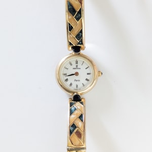 May include: A gold-toned Festina Paris watch with a white face and Roman numeral hour markers. The watch band features a woven design with black and teal accents. The watch has a small gold crown and a black accent on the band.