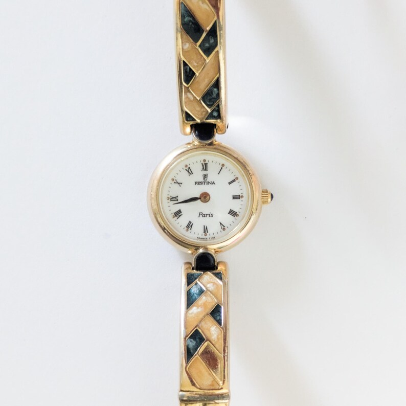 May include: A gold-toned Festina Paris watch with a white face and Roman numeral hour markers. The watch band features a woven design with black and teal accents. The watch has a small gold crown and a black accent on the band.
