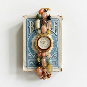 May include: A gold-toned wristwatch with a white face and a beaded strap. The strap is made of various colorful gemstones, including orange, green, and black beads. The watch is resting on a vintage blue and white playing card box.