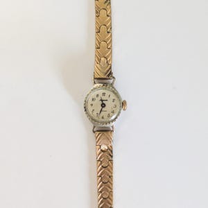 May include: A vintage gold-toned wristwatch with a round, silver-colored face. The watch features black numerals and hands, with a gold-toned, textured band. The watch face reads "Ingersoll" and "Phoenix".