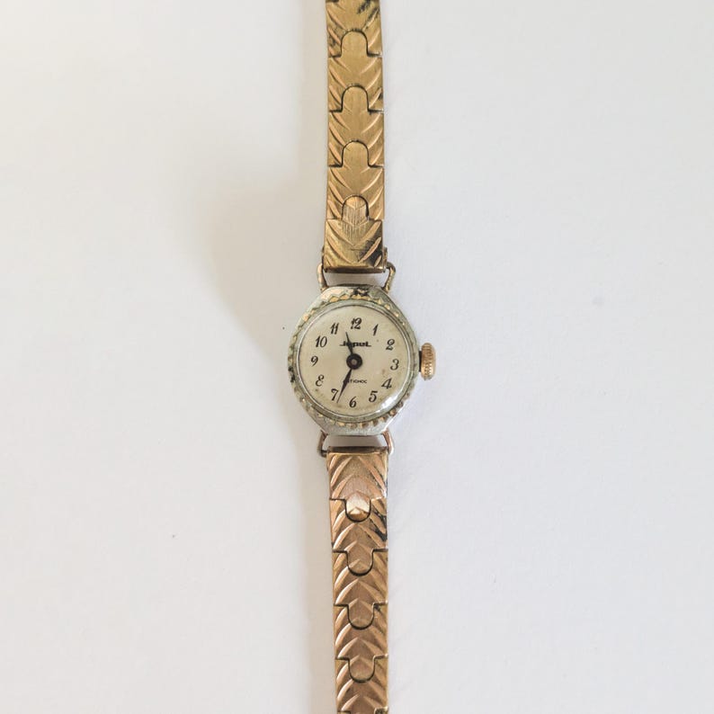 May include: A vintage gold-toned wristwatch with a round, silver-colored face. The watch features black numerals and hands, with a gold-toned, textured band. The watch face reads "Ingersoll" and "Phoenix".