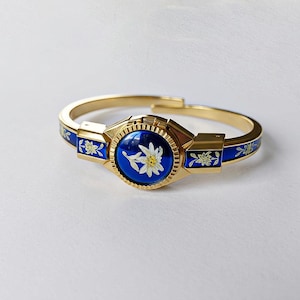 Può includere: Un bracciale rigido dorato con dettagli in smalto blu. Il bracciale presenta un medaglione centrale blu con un motivo floreale bianco e giallo. Motivi floreali adornano le sezioni in smalto blu. Il bracciale ha una chiusura.