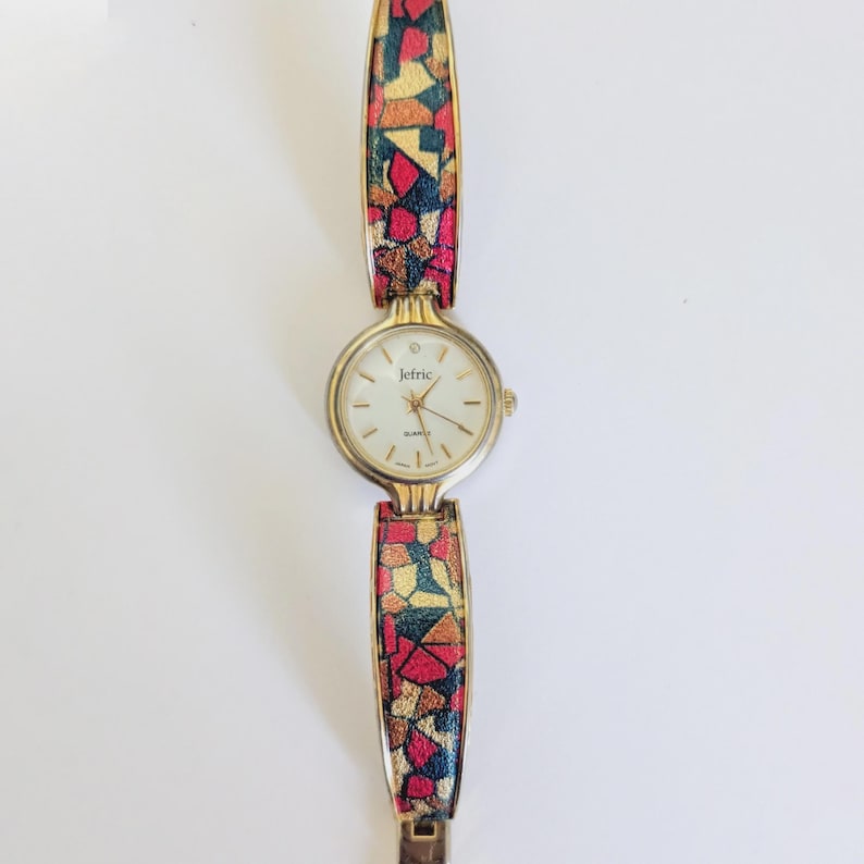 May include: A gold-toned Jetric quartz watch with a white face and gold hands. The watch band features a colorful mosaic pattern in red, yellow, and green. The watch is displayed on a white surface.