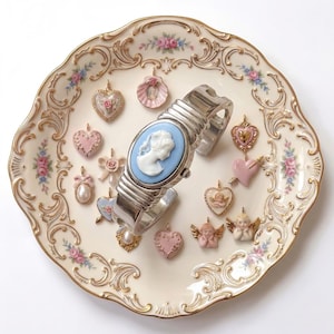 May include: A collection of vintage-style jewelry and a watch on a decorative plate. The watch features a blue cameo face and a silver band. Various heart-shaped and cherub-themed charms in pink and gold surround the watch.