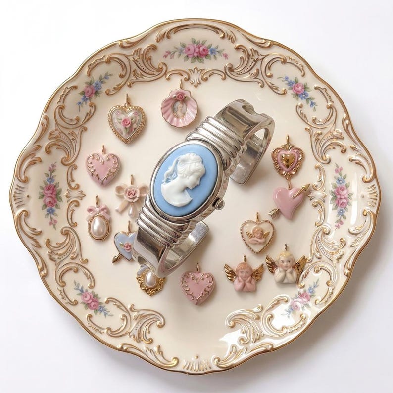 May include: A collection of vintage-style jewelry and a watch on a decorative plate. The watch features a blue cameo face and a silver band. Various heart-shaped and cherub-themed charms in pink and gold surround the watch.