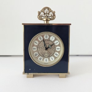 Puede incluir: Un reloj de viaje Europa vintage con una caja rectangular azul marino y una parte superior decorativa dorada. La esfera del reloj es de color crema con números romanos y un centro decorativo. El reloj tiene pies dorados.