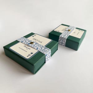 May include: Two dark green gift boxes with decorative Ace of Spades playing card labels. Each box is wrapped with a patterned ribbon and a gold button. The labels read "TICK & Trick" and "Thank you".