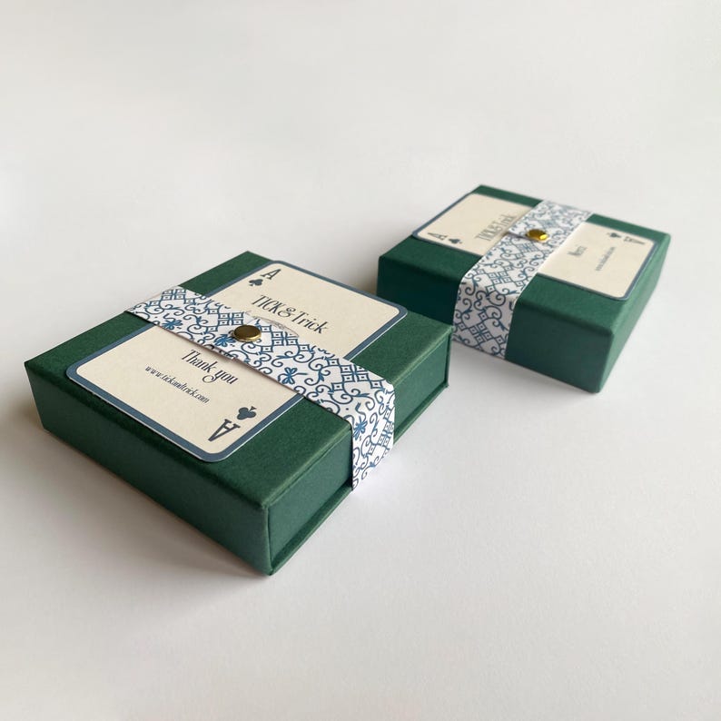 May include: Two dark green gift boxes with decorative Ace of Spades playing card labels. Each box is wrapped with a patterned ribbon and a gold button. The labels read "TICK & Trick" and "Thank you".