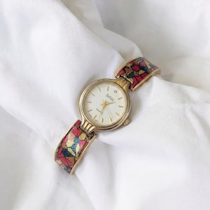 May include: A gold-toned Jefric quartz watch with a white face and a single diamond accent. The watch band features a colorful mosaic pattern in red, yellow, and blue. The watch is set against a white background.