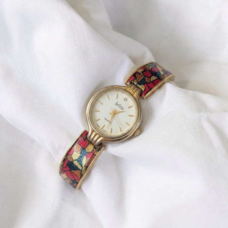 May include: A gold-toned Jefric quartz watch with a white face and a single diamond accent. The watch band features a colorful mosaic pattern in red, yellow, and blue. The watch is set against a white background.