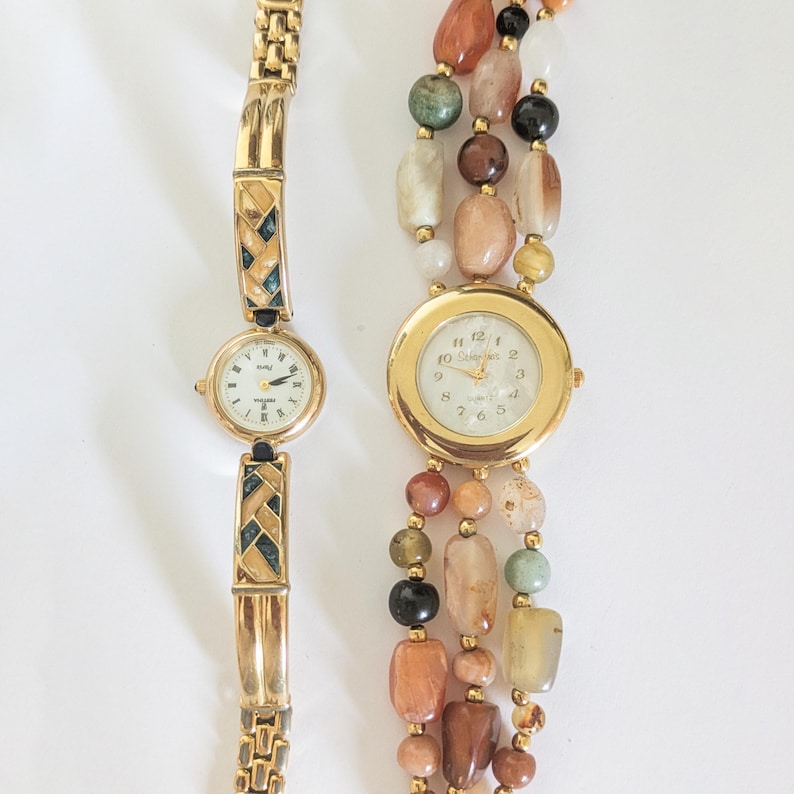 May include: Two vintage women's watches. One has a gold-tone band with geometric accents and a white face. The other watch has a gold-tone face and is surrounded by strands of colorful beads.