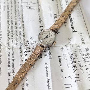 May include: A vintage gold-toned wristwatch with a small, round face and a textured band. The watch rests on a document with French text, showcasing its classic design and intricate details. The watch face has a diameter of approximately 1 inch.