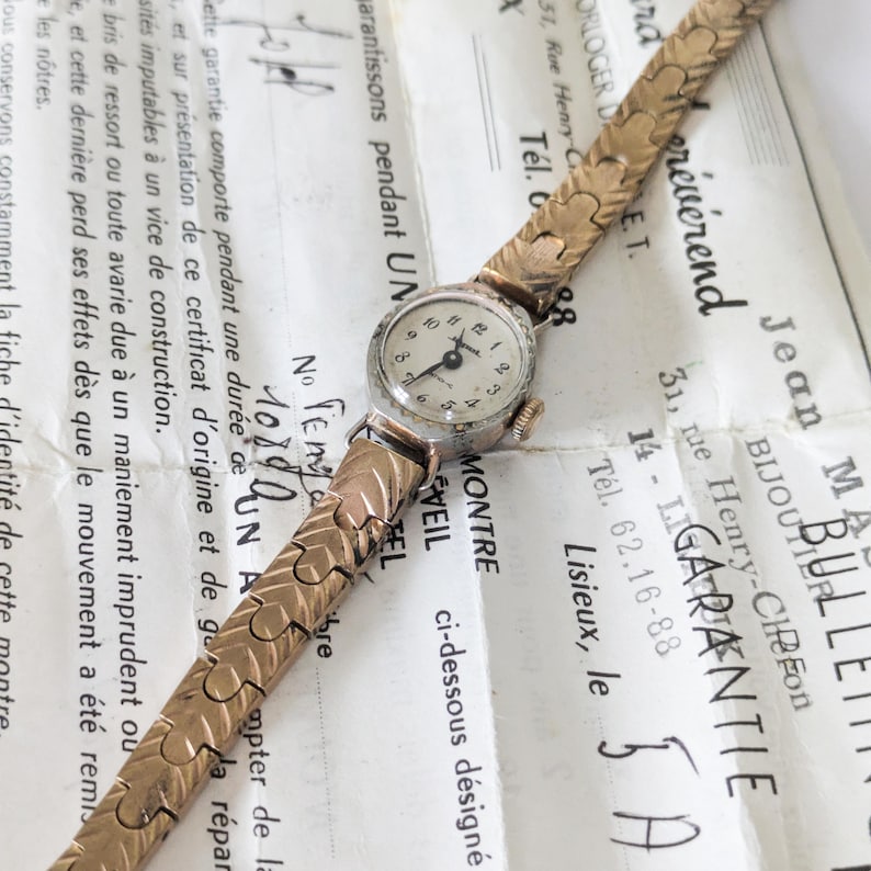 May include: A vintage gold-toned wristwatch with a small, round face and a textured band. The watch rests on a document with French text, showcasing its classic design and intricate details. The watch face has a diameter of approximately 1 inch.