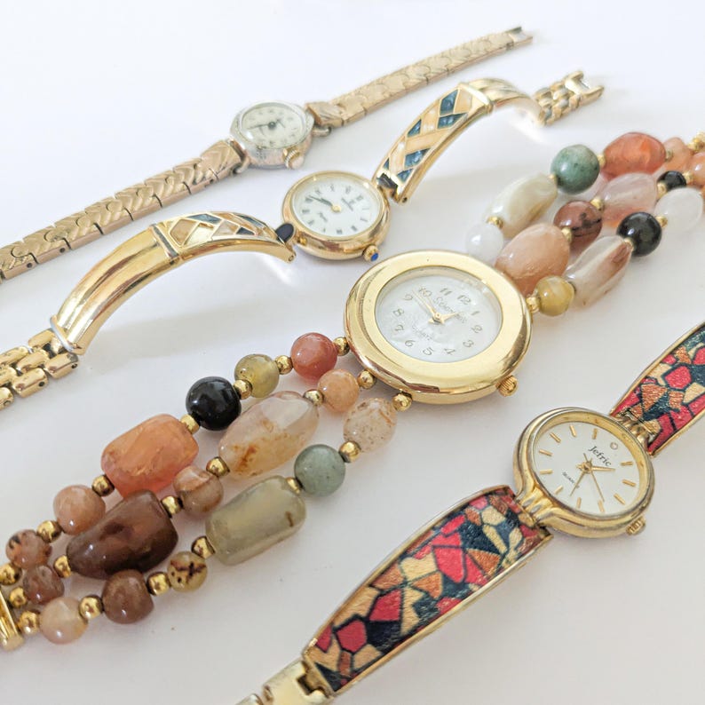 May include: A collection of vintage women's watches with gold-toned metal bands and watch faces. One watch features a beaded band with earth-toned beads. Other bands have geometric and textured designs.