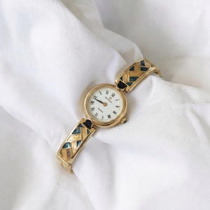 May include: Gold-toned wristwatch with a white face and black Roman numerals. The watch band features a geometric pattern with teal and black accents. The watch is displayed on a white, textured surface.
