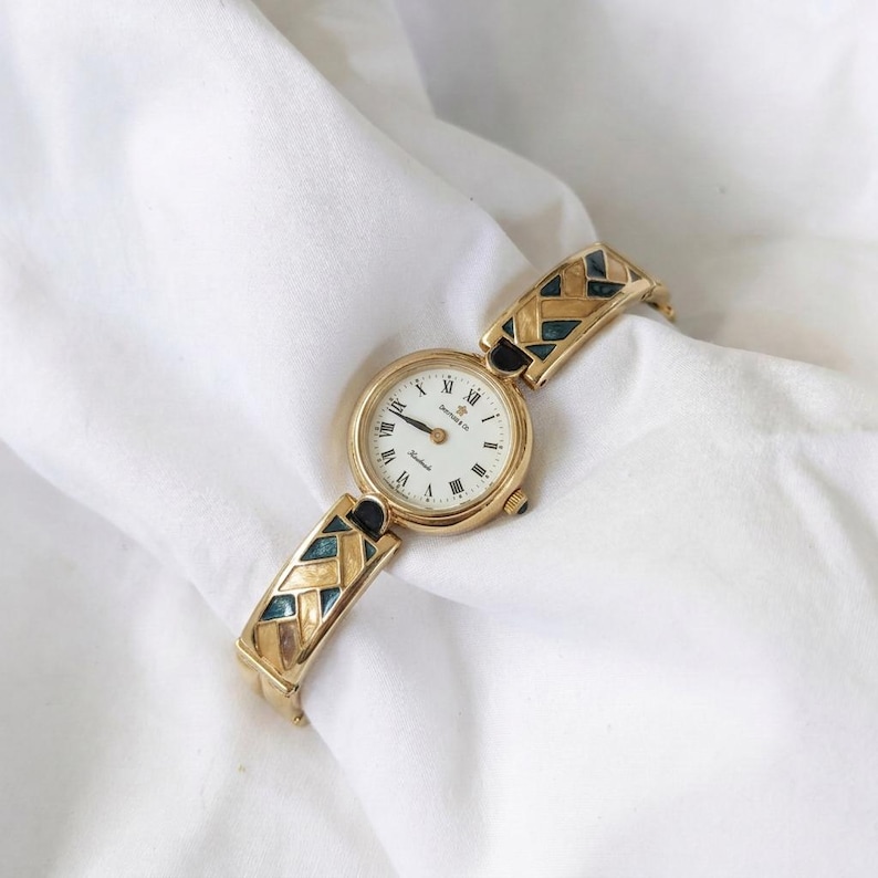 May include: Gold-toned wristwatch with a white face and black Roman numerals. The watch band features a geometric pattern with teal and black accents. The watch is displayed on a white, textured surface.
