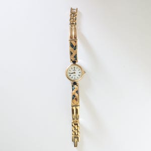 May include: A gold-toned wristwatch with a round, white face and black hands. The watch band features a woven design with dark blue accents. The watch is displayed on a white surface.
