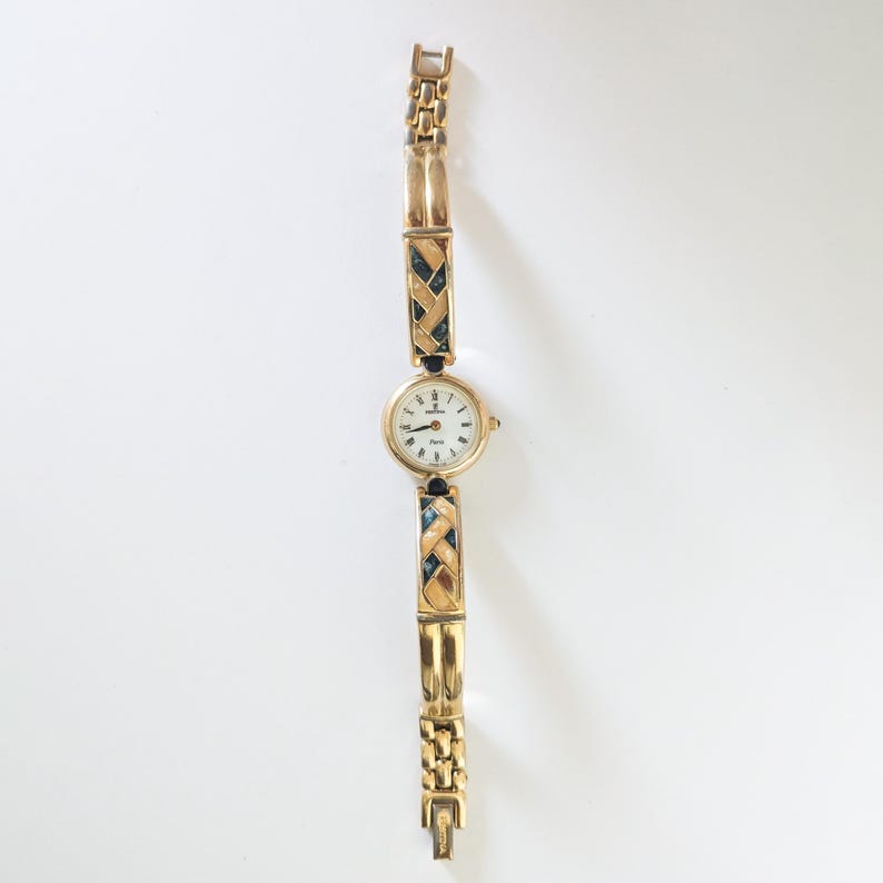 May include: A gold-toned wristwatch with a round, white face and black hands. The watch band features a woven design with dark blue accents. The watch is displayed on a white surface.