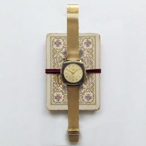 Peut inclure: Une montre-bracelet dorée avec un bracelet en maille et un cadran carré. La montre est posée sur une carte vintage à motifs floraux, maintenue par un ruban bordeaux. Le cadran de la montre porte le mot "GAB".