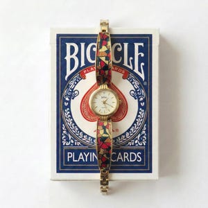 May include: A gold-toned wristwatch with a patterned band in red, yellow, and black, resting on a Bicycle playing cards box. The watch face is white with gold hands and the brand name "Jetric". The playing cards box has a blue and white design.