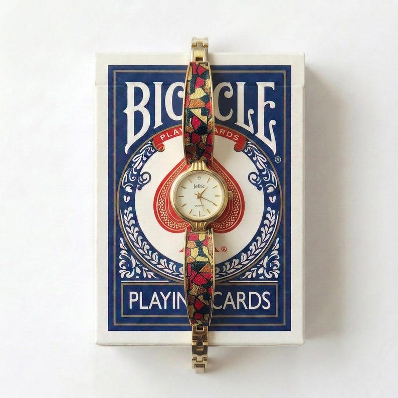 May include: A gold-toned wristwatch with a patterned band in red, yellow, and black, resting on a Bicycle playing cards box. The watch face is white with gold hands and the brand name "Jetric". The playing cards box has a blue and white design.