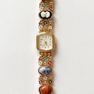 Peut inclure: Une montre-bracelet dorée avec un cadran rectangulaire et un bracelet décoratif. Le bracelet présente divers ornements colorés et ornés, dont un camée noir, une pierre bleue et des accents rouges. Le cadran affiche les chiffres de 1 à 12.