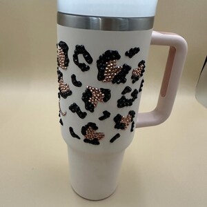 May include: A white insulated tumbler with a pink handle and lid. The tumbler is decorated with black and rose gold leopard print accents. The lid is white and the handle is a soft pink color. The tumbler is designed to keep beverages hot or cold.
