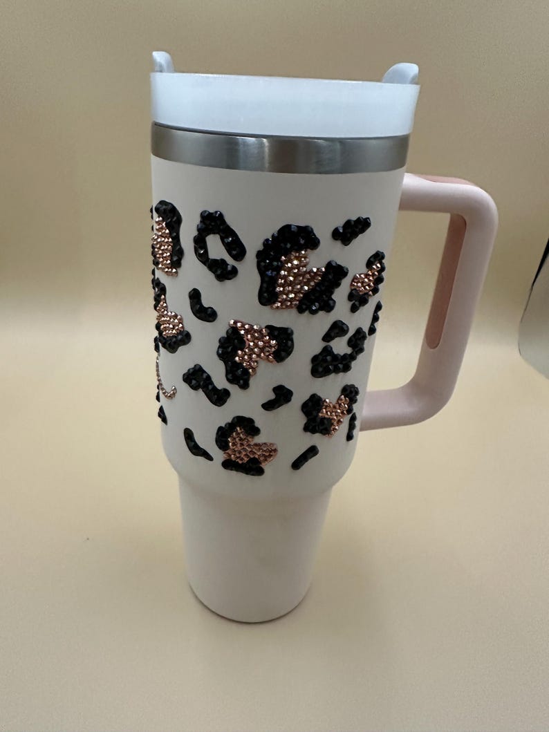 May include: A white insulated tumbler with a pink handle and lid. The tumbler is decorated with black and rose gold leopard print accents. The lid is white and the handle is a soft pink color. The tumbler is designed to keep beverages hot or cold.