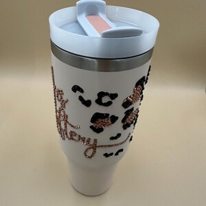 May include: White tumbler with a white lid and a silver band. The tumbler features black leopard print spots and the word "Merry" in rose gold glitter. The lid has a sliding closure with a peach-colored accent.