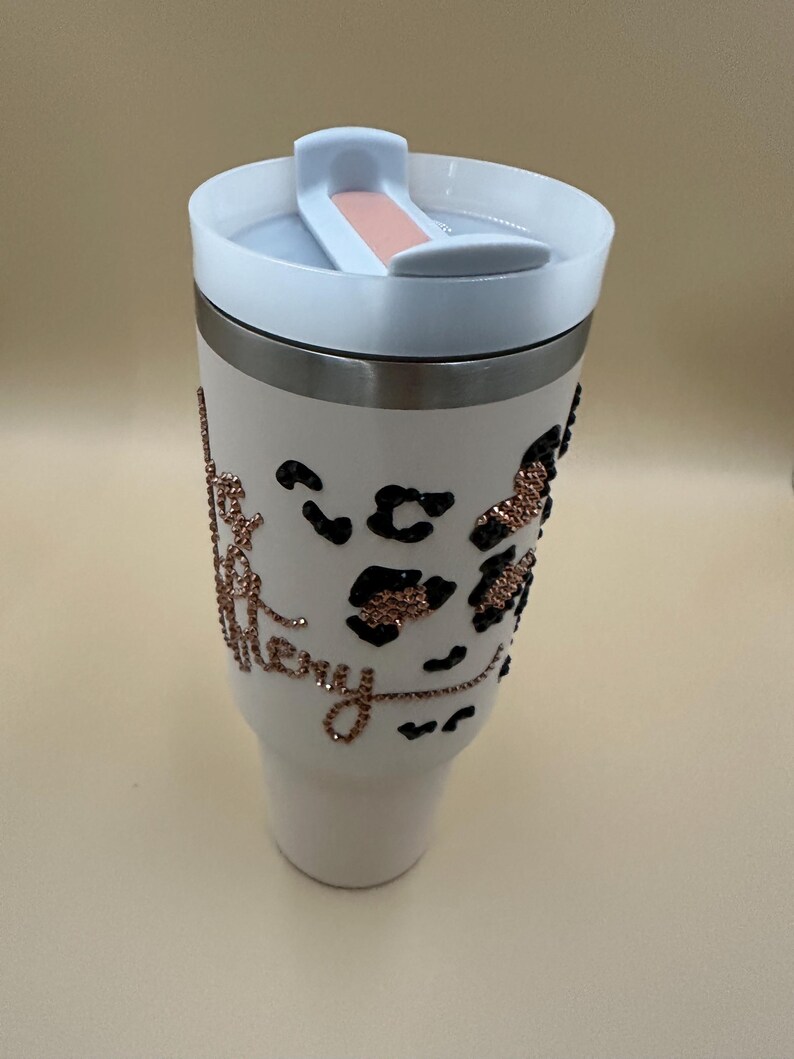 May include: White tumbler with a white lid and a silver band. The tumbler features black leopard print spots and the word "Merry" in rose gold glitter. The lid has a sliding closure with a peach-colored accent.