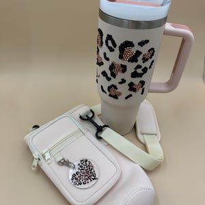 May include: A light pink insulated tumbler with a handle, decorated with black and rose gold leopard print accents. Includes a matching light pink water bottle carrier with a shoulder strap and a leopard print heart-shaped charm.