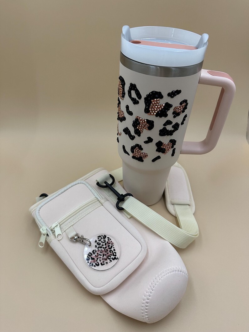 May include: A light pink insulated tumbler with a handle, decorated with black and rose gold leopard print accents. Includes a matching light pink water bottle carrier with a shoulder strap and a leopard print heart-shaped charm.