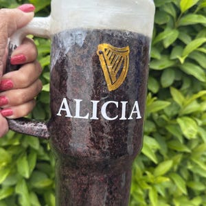 Pu&ograve; includere: Un bicchiere personalizzato con un esterno marrone scuro e glitterato, simile a una pinta di stout. Il bicchiere presenta il nome "ALICIA" in lettere bianche, un logo dell'arpa dorata e una parte superiore bianca simile alla schiuma. Include una cannuccia.