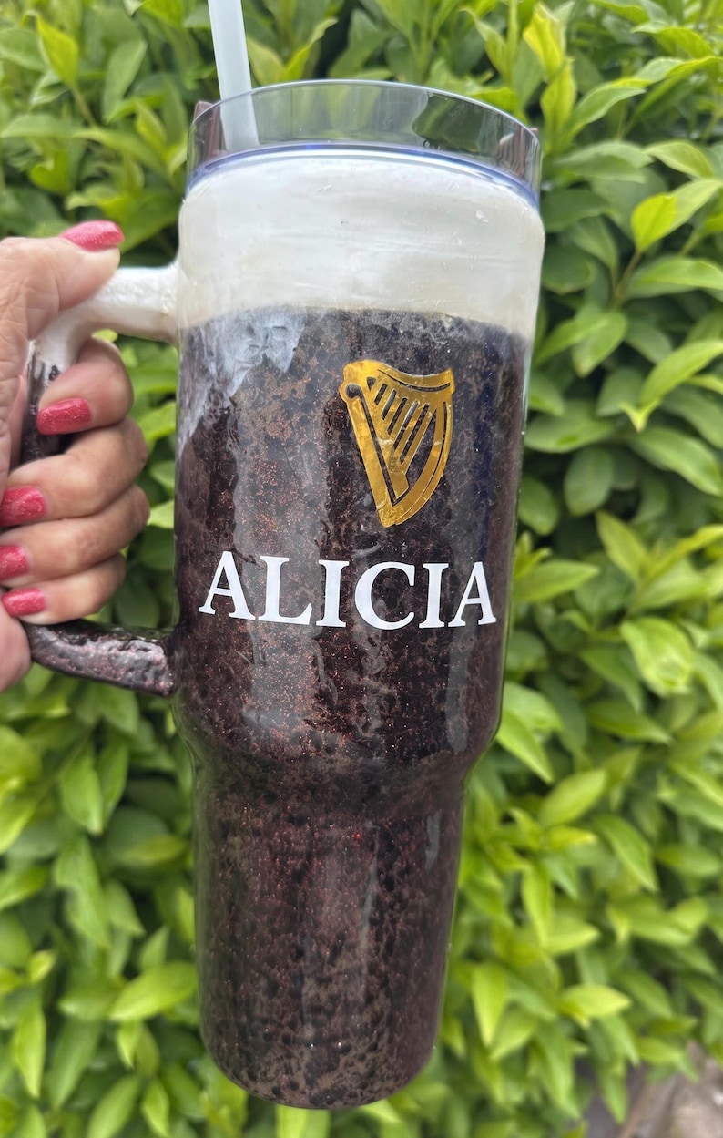 Pu&ograve; includere: Un bicchiere personalizzato con un esterno marrone scuro e glitterato, simile a una pinta di stout. Il bicchiere presenta il nome "ALICIA" in lettere bianche, un logo dell'arpa dorata e una parte superiore bianca simile alla schiuma. Include una cannuccia.