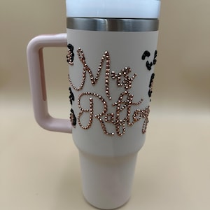May include: A white insulated tumbler with a pink handle and lid. The tumbler is adorned with rose gold rhinestones spelling out "Mrs. Raftery" and black leopard print accents. The lid is white and the rim is silver.