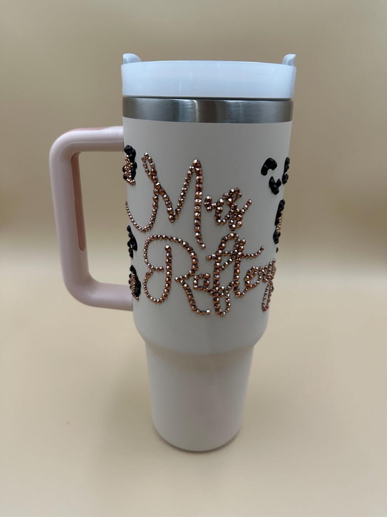 May include: A white insulated tumbler with a pink handle and lid. The tumbler is adorned with rose gold rhinestones spelling out "Mrs. Raftery" and black leopard print accents. The lid is white and the rim is silver.
