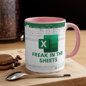May include: White ceramic mug with a pink interior and handle. The mug features a design resembling a spreadsheet, with the Microsoft Excel logo and the text "FREAK IN THE SHEETS" in green.