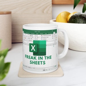 May include: White ceramic mug featuring an Excel spreadsheet design with the Microsoft Excel logo and the text "FREAK IN THE SHEETS" in green. The mug has a white handle and sits on a beige coaster.