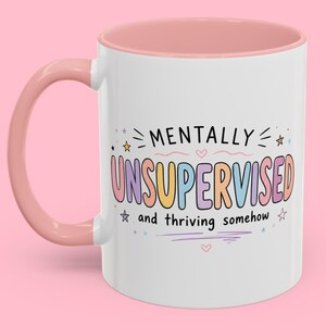 Pode incluir: Caneca de cerâmica branca com alça e interior rosa. A caneca apresenta o texto "MENTALLY UNSUPERVISED and thriving somehow" em letras coloridas e divertidas, com estrelas e corações.