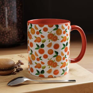 May include: White ceramic mug with a red interior and handle. The mug is decorated with a pattern of oranges, orange slices, and green leaves. The mug is sitting on a wooden surface with a spoon and a pastry.