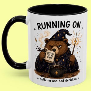 Könnte beinhalten: Weiße Keramiktasse mit schwarzem Henkel und Rand. Die Tasse zeigt einen Cartoon-Bären als Zauberer, der eine Kaffeetasse mit der Aufschrift "Caffeine Fueling Chaos" hält. Die Tasse trägt auch den Text "Running on caffeine and bad decisions."