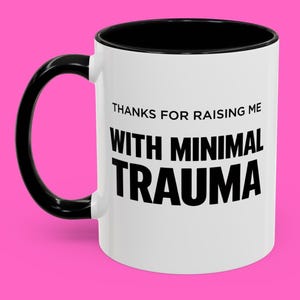 Könnte beinhalten: Weißer Keramikbecher mit schwarzem Henkel und Innenraum. Der Becher trägt den Text "THANKS FOR RAISING ME WITH MINIMAL TRAUMA" in schwarzer, fetter Schrift. Der Becher steht vor einem hellrosa Hintergrund.