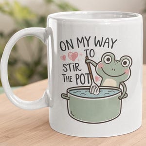 May include: White ceramic mug with a green frog illustration stirring a pot of blue liquid. The mug has the text "On my way to stir the pot" with pink hearts.