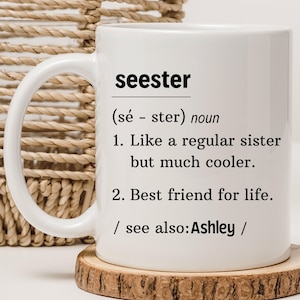 以下が含まれることがあります： 白磁のマグカップに、「seester」という言葉の白黒の定義が書かれています。「普通の妹みたいだけど、もっとクール」と「一生の親友」と定義されています。定義には、「参考：Ashley」というフレーズも含まれています。