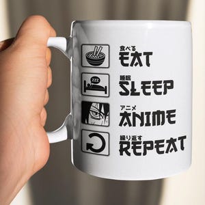 May include: White ceramic mug with black text and illustrations. The text reads "Eat, Sleep, Anime, Repeat" in English and Japanese. The illustrations include a bowl of ramen, a bed, an anime character's eye, and a circular arrow.