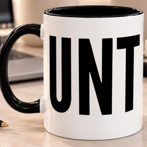 May include: A white ceramic mug with a black interior and a black handle. The exterior features the word CUNT printed in large, bold, black capital letters. The design is simple and high-contrast, with the text wrapping around the front surface.