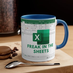 May include: White ceramic mug with a blue interior and handle. The mug features an Excel spreadsheet design with the text "FREAK IN THE SHEETS" in green. The mug is on a wooden surface with a spoon and a pastry.