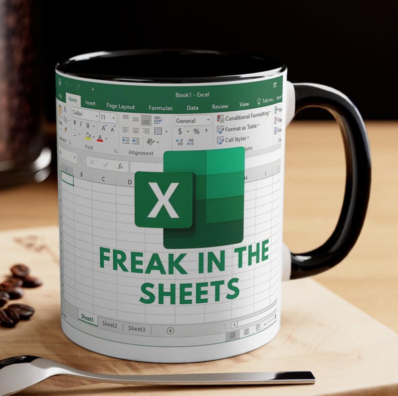 May include: White ceramic mug with a black interior and handle. The mug features an Excel spreadsheet design with the text "FREAK IN THE SHEETS" and the Excel logo. A spoon and coffee beans are in the foreground.