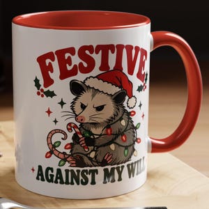 Op de afbeelding: Een witte keramische mok met een rode binnenkant en handvat, voorzien van een afbeelding van een chagrijnige opossum met een kerstmuts, gewikkeld in kleurrijke kerstverlichting en een zuurstok vasthoudend, met de tekst FESTIVE AGAINST MY WILL.